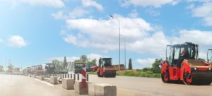 Several construction vehicles are parked on an asphalt road, in daytime, with a light blue sky overhead. A worker in safety clothing can be seen near the machines.